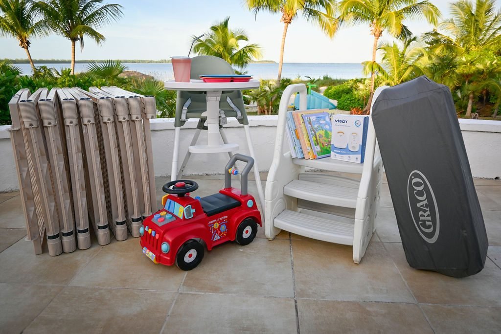 Children's equipment at Hightide Estate — high chair, ride-on toys, books, pack-and-play crib, and baby gates on ocean-view patio